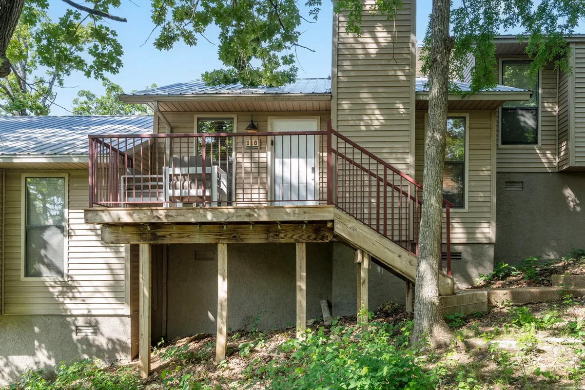 The image features an inviting deck with a railing, the entrance door to the property, and mature trees surrounding the space. The lighting suggests a warm, welcoming atmosphere, ideal for relaxing or gathering outdoors.