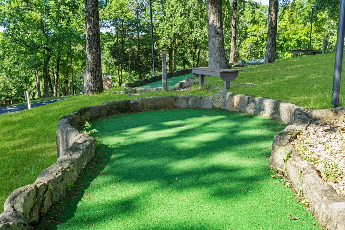 The mini-golf putting green surrounded by mature trees and a charming stone border. The area is set up for friendly competitions and group activities, offering a playful, family-friendly vibe.