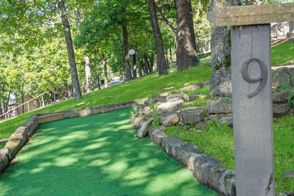 This image features a vibrant mini-golf hole surrounded by lush green synthetic turf, bordered with stones, and shaded by mature trees. The setting is outdoors, evoking a sense of playful competition and family fun, with plenty of space for multiple players to gather and enjoy the game together.