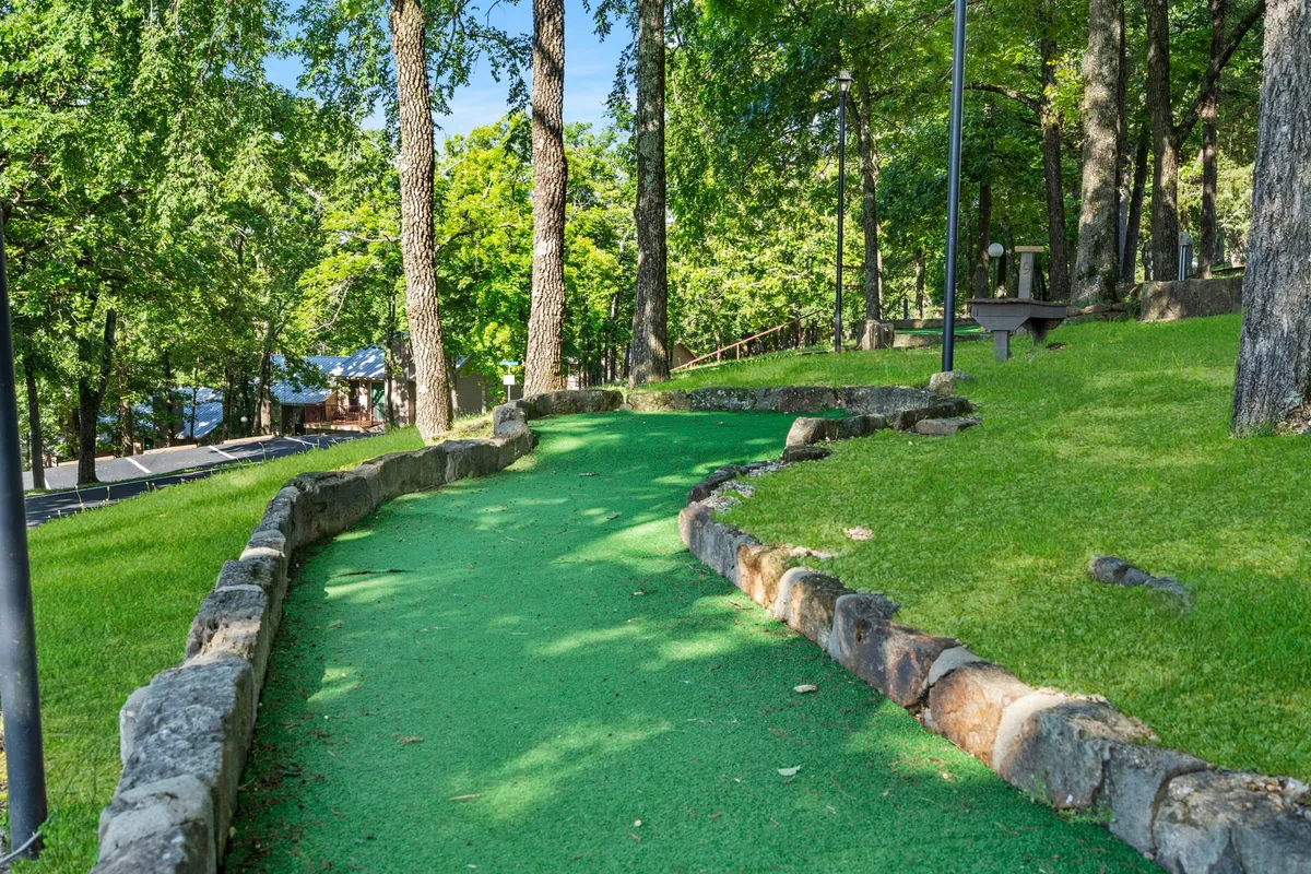 A playful, inviting mini-golf course winding through natural surroundings with mature trees and rock borders. The green pathway hints at group and family fun, with the setting blending seamlessly into the forested landscape.