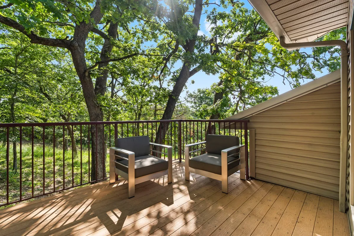 A tranquil front porch featuring two modern chairs on a wooden deck surrounded by lush greenery. Soft morning or evening light filters through the trees, creating a peaceful and inviting outdoor space perfect for relaxing with coffee or keeping a pet companion nearby.