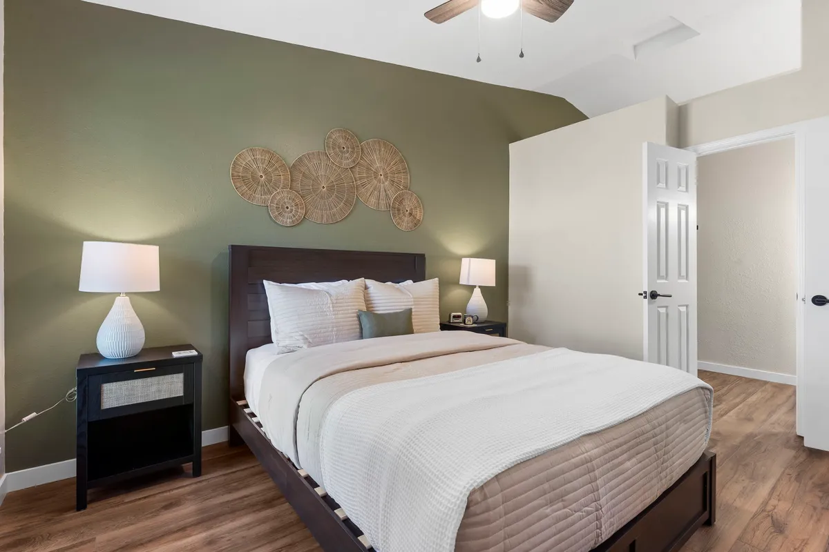 A tranquil bedroom featuring a plush bed adorned with decorative pillows, woven wall art above, and a nightstand with a softly glowing lamp. Sunlight streams in, creating a peaceful ambiance ideal for relaxation.