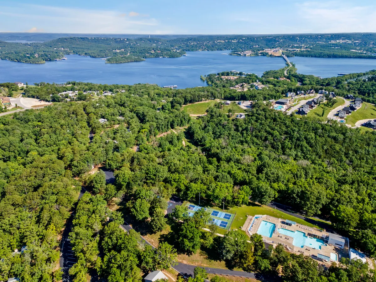 A sweeping exterior view showcasing a scenic lake, a shimmering swimming pool, and adjacent tennis courts nestled amidst lush greenery. The tranquil environment is complemented by hints of walking trails and recreational spaces, emphasizing both water and land activities available at the resort.