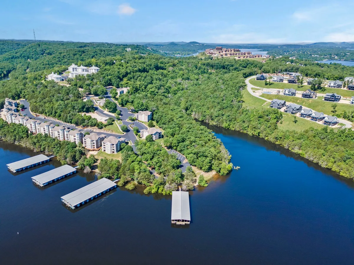 A tranquil waterfront area with green hills in the background, residential buildings along the shore, and marinas filled with boats. People can be seen boating, fishing, and relaxing by the water as the sun begins to set, casting a golden glow over the scene.