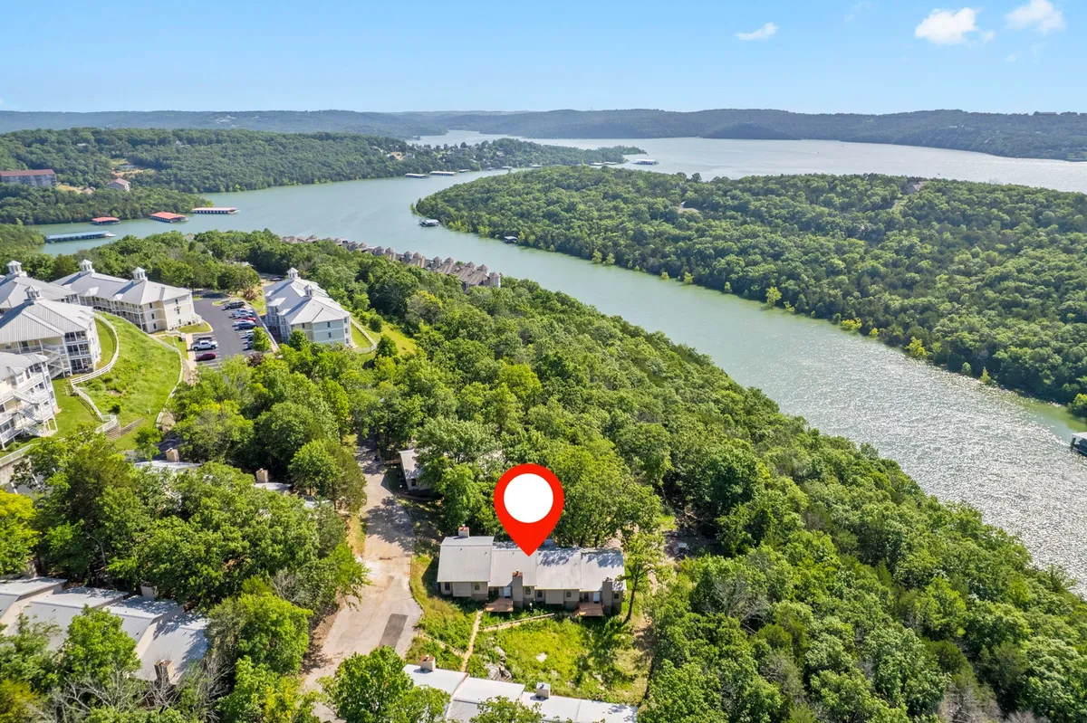 A scenic view of the house nestled among trees with a river and hills in the background, capturing the essence of the Ozarks. The landscape invites outdoor activity such as walking, relaxing by the water, or taking in wildlife at sunset.